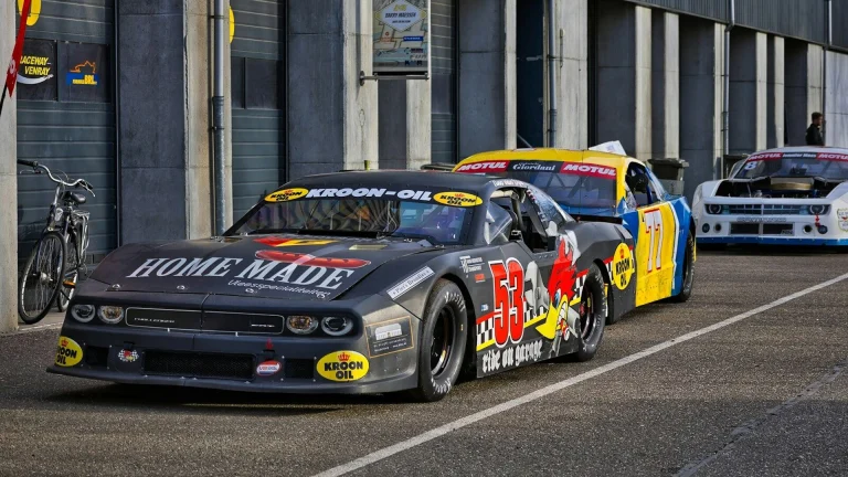 Race cars lined up on track with pit lane backdrop.