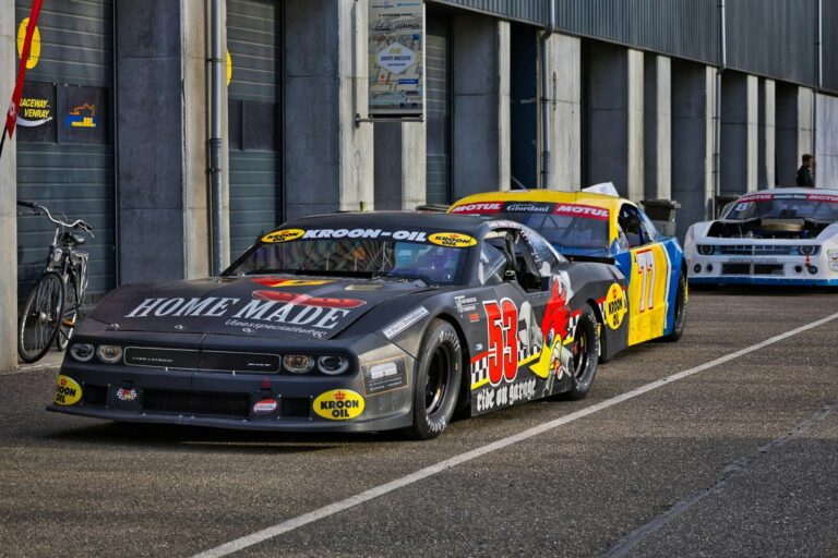 Race cars lined up on track with pit lane backdrop.