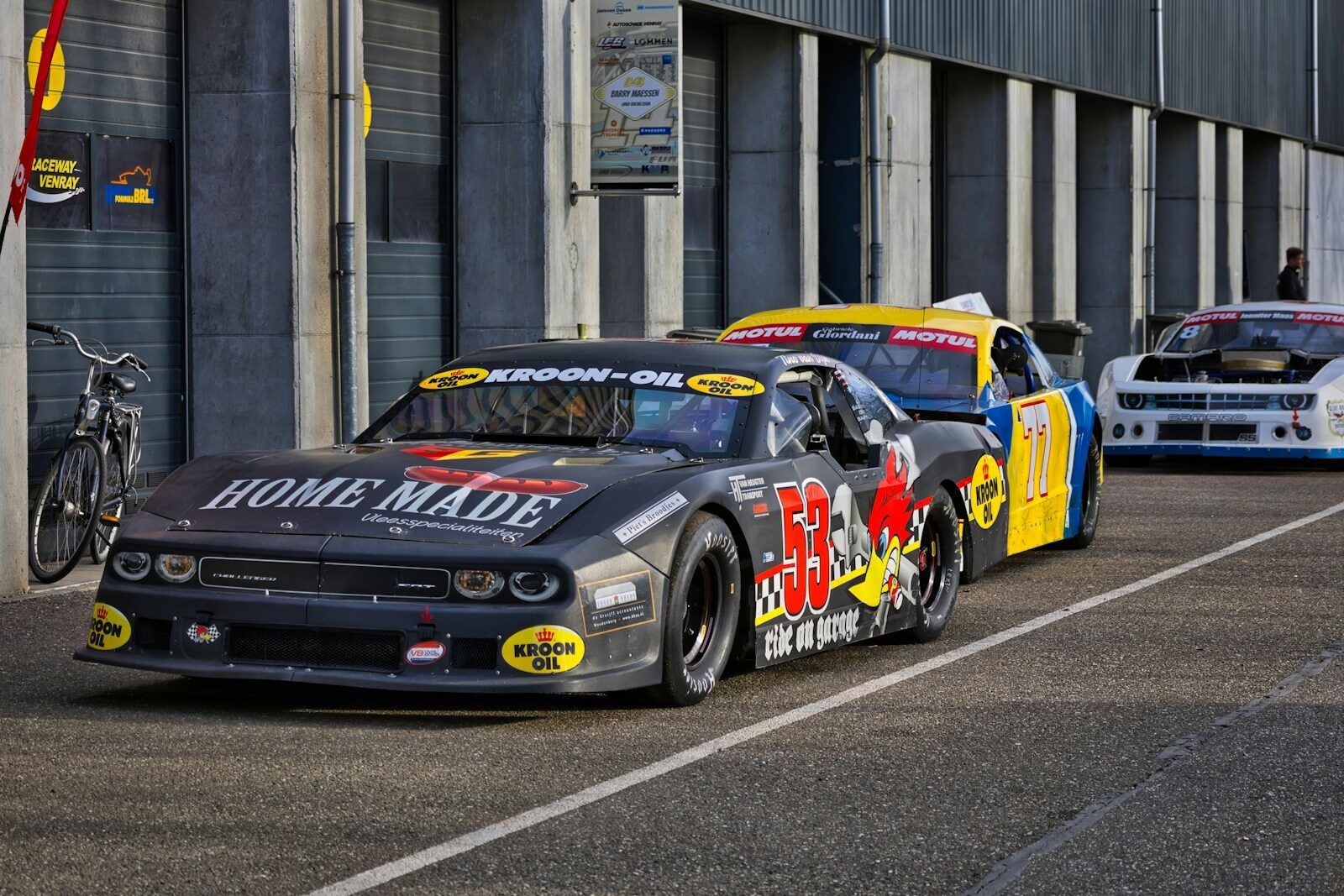 Race cars lined up on track with pit lane backdrop.