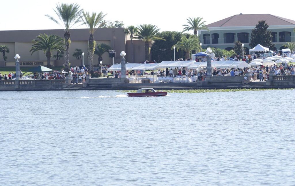 A red Amphicar 770 crossing a lake past a crowd of onlookers at a classic car show