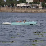 A mint-green Amphicar 770 convertible driving through open water, kicking up a wake behind it
