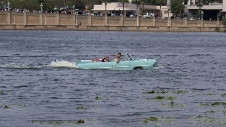 A mint-green Amphicar 770 convertible driving through open water, kicking up a wake behind it