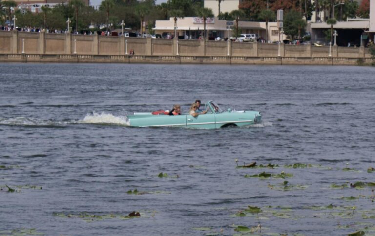 A mint-green Amphicar 770 convertible driving through open water, kicking up a wake behind it