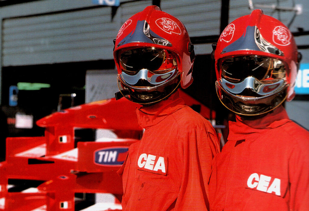 Two CEA Squadra Corse track firefighters in red suits and red helmets with CEA lettering