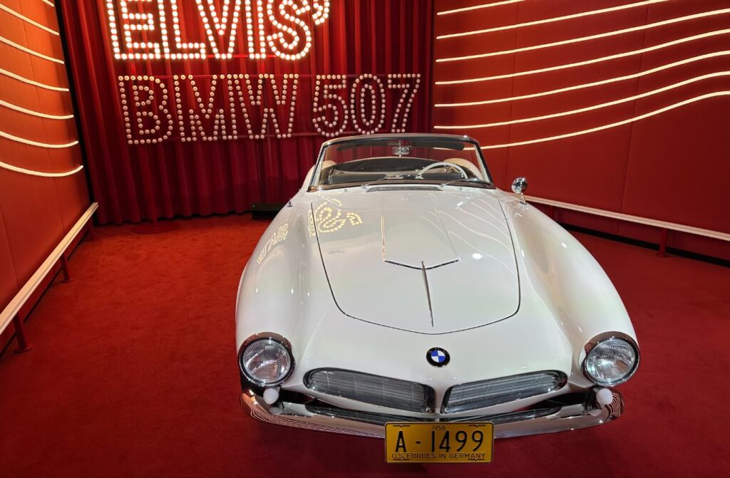 Elvis Presley's restored BMW 507, repainted back to white, on display beneath an "Elvis' BMW 507" sign with a replica U.S. Forces in Germany military plate