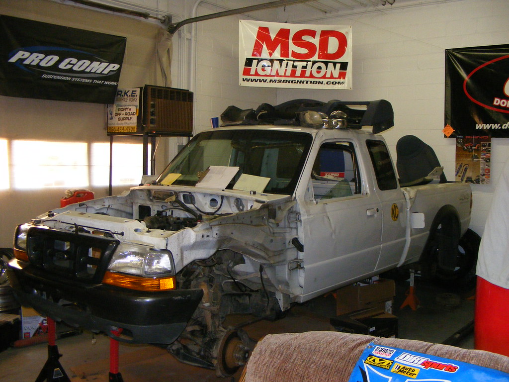 A silver pickup truck with its front end stripped down on jack stands in a garage