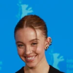 Sydney Sweeney smiling in a black blazer against a blue Berlinale backdrop