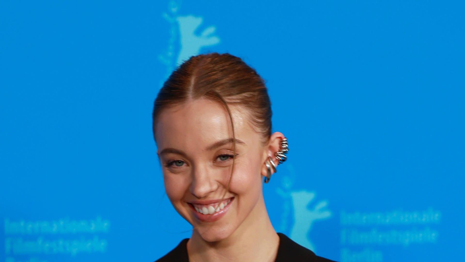 Sydney Sweeney smiling in a black blazer against a blue Berlinale backdrop