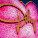 A large pink sequined heart sign with gold cursive lettering against a blue sky