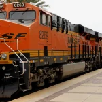 An orange BNSF locomotive pulling a long string of intermodal container cars past a station platform