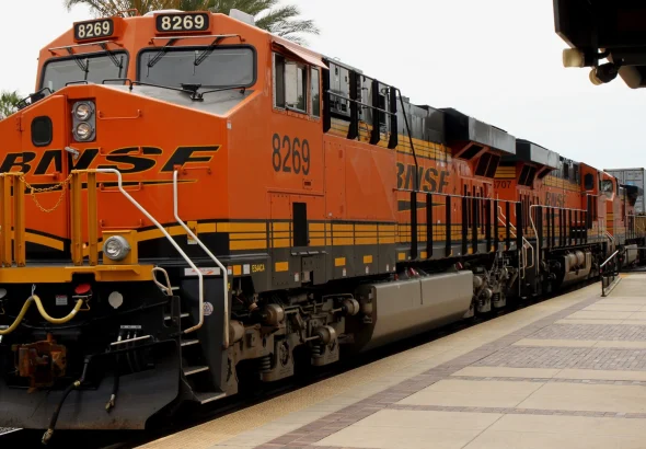An orange BNSF locomotive pulling a long string of intermodal container cars past a station platform