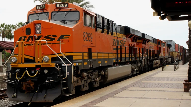 An orange BNSF locomotive pulling a long string of intermodal container cars past a station platform