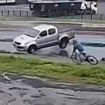 Surveillance video still of a silver Toyota Hilux pickup sliding toward a cyclist on a wet service road beside BR-163 in Marechal Cândido Rondon, Brazil