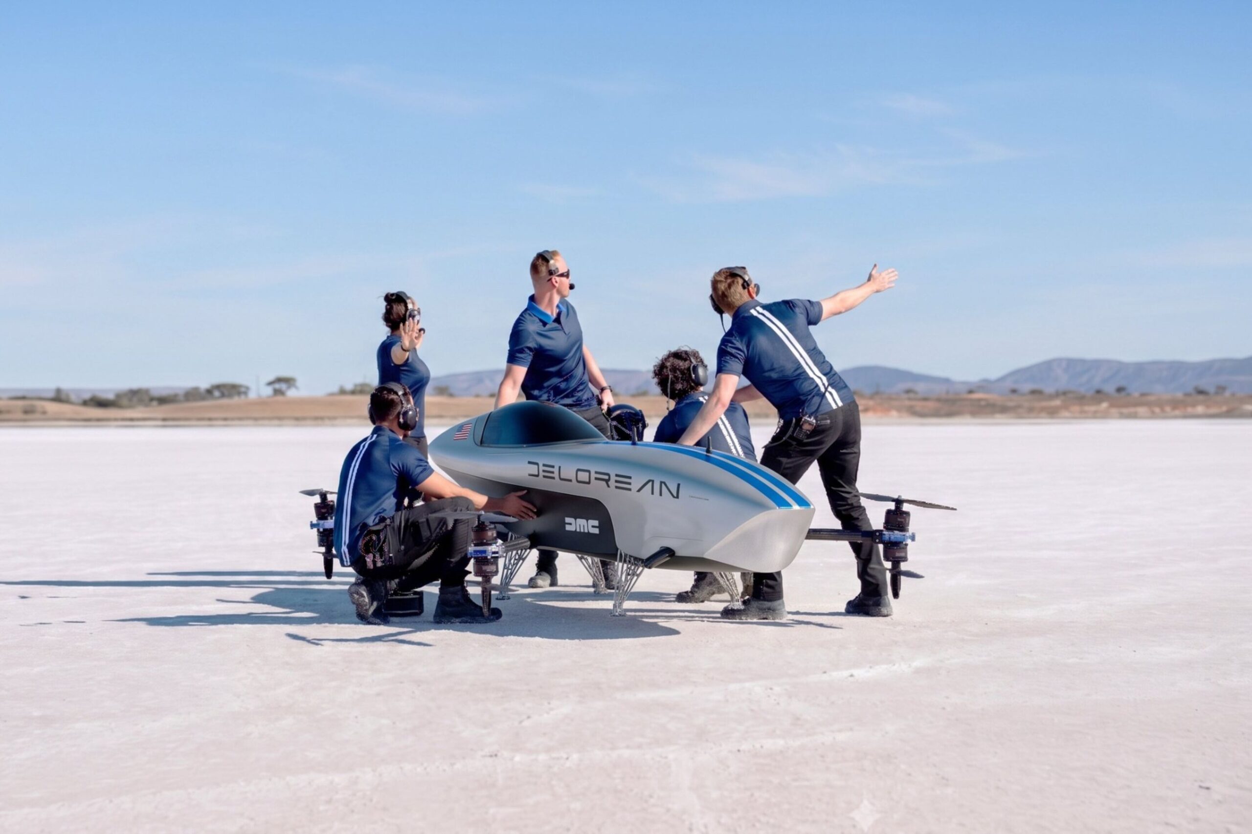 Four crew members in navy shirts working around a silver DeLorean-liveried Airspeeder aircraft on a flat white surface
