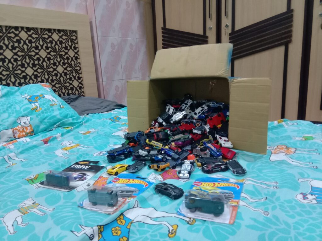 A cardboard box tipped over with dozens of Hot Wheels die-cast cars spilling across a bedspread