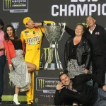 Kyle Busch in his yellow firesuit beside the trophy on the 2019 NASCAR Cup Series champion stage