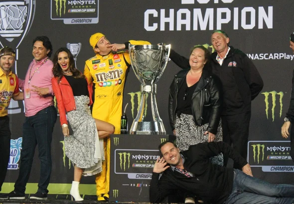 Kyle Busch in his yellow firesuit beside the trophy on the 2019 NASCAR Cup Series champion stage