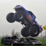Blue Bigfoot monster truck landing on a row of junked cars