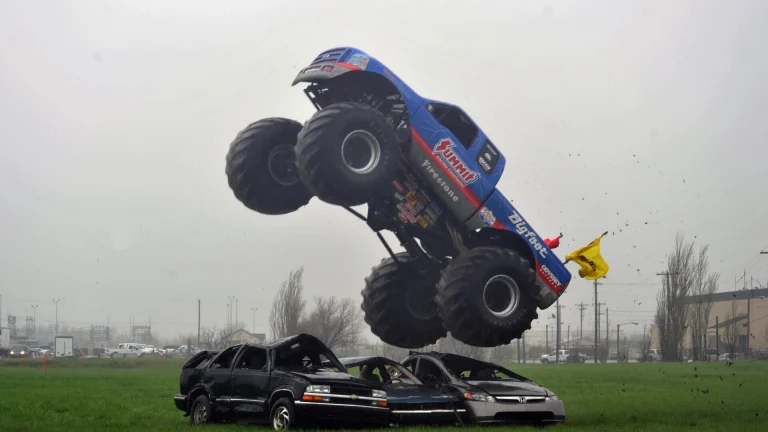 Blue Bigfoot monster truck landing on a row of junked cars