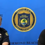 Two uniformed Ormond Beach Police Department officers seated in front of a department backdrop