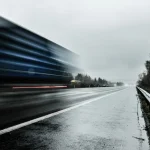 A semi-truck blurred by speed on a wet, overcast interstate beside a guardrail