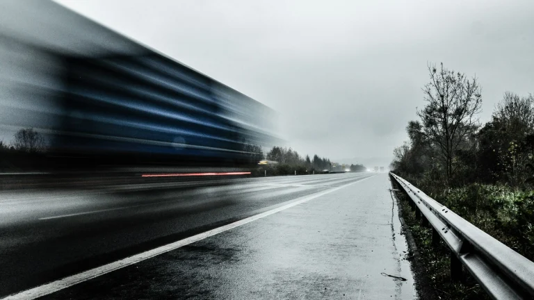 A semi-truck blurred by speed on a wet, overcast interstate beside a guardrail